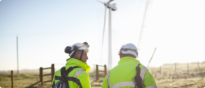 Two men in yellow vests stand beside a wind turbine, highlighting renewable energy efforts in a clear, sunny environment.