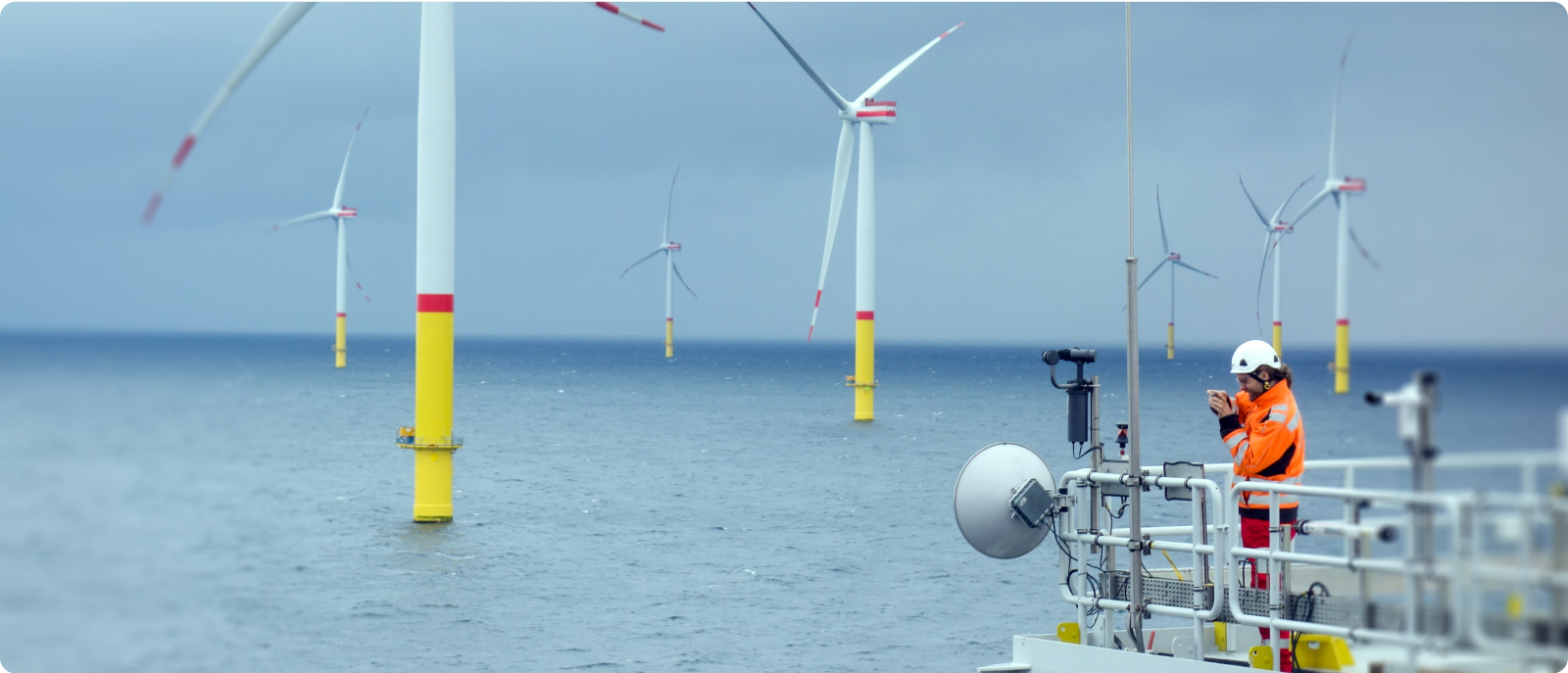 On a boat deck, a man wearing an orange vest observes wind turbines in the distance, highlighting renewable energy efforts.