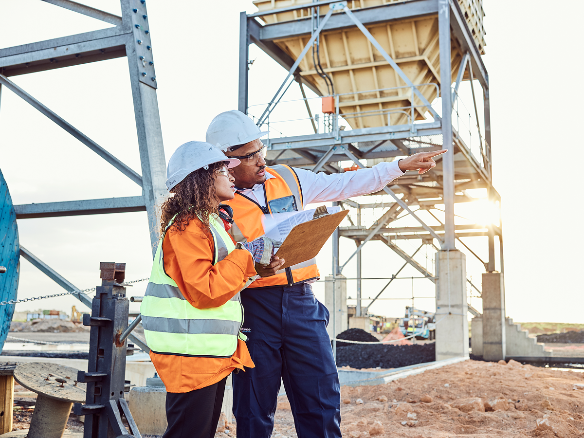 Two individuals in hard hats and safety vests stand before a construction site, discussing the ongoing project.