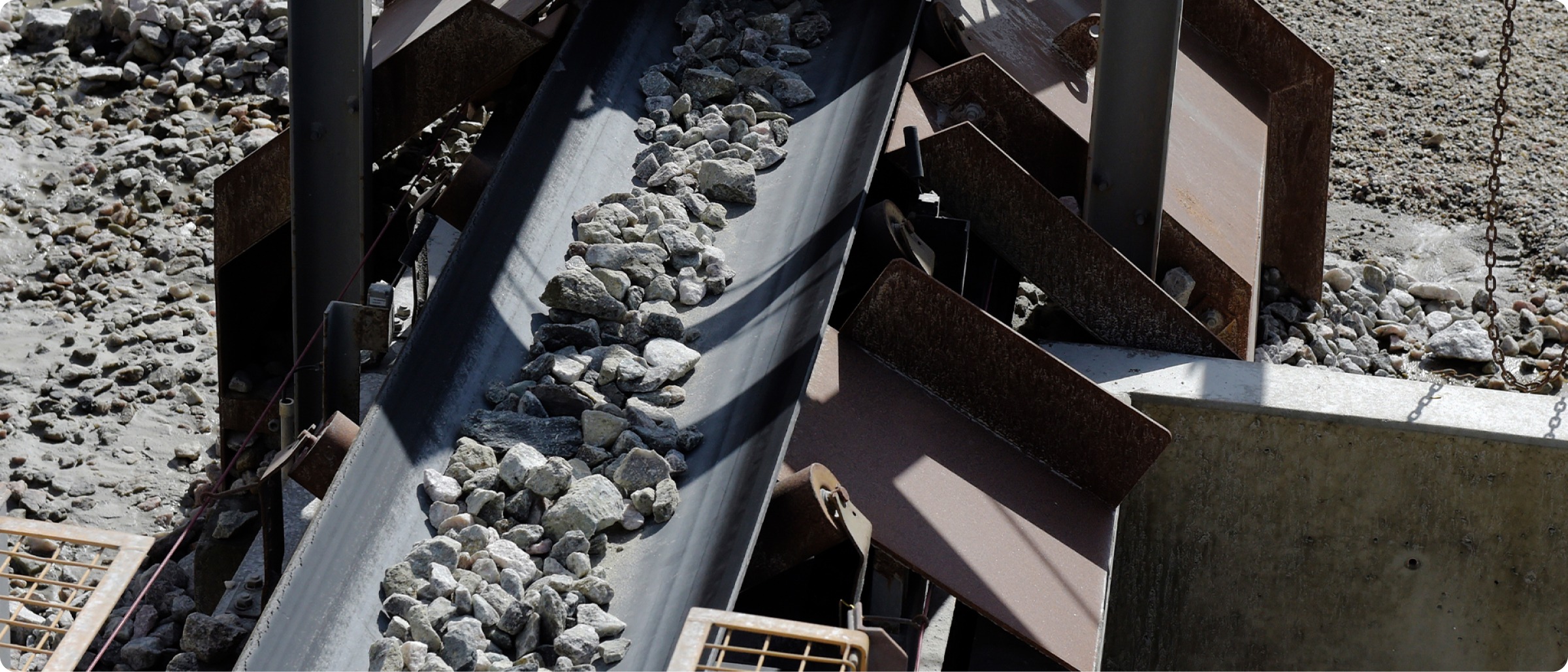 A conveyor belt transports a load of small gray rocks through an industrial processing system, surrounded by metal supports and machinery components.
