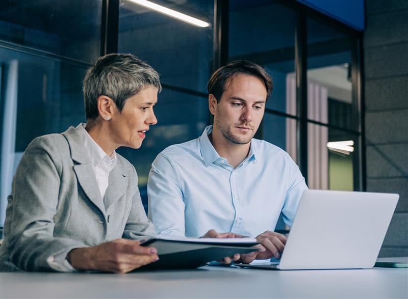 Two people sitting at a desk in an office setting, reviewing documents together while working on a laptop.