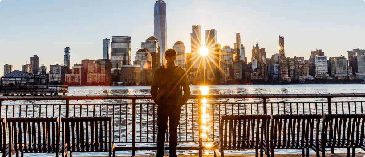 A man stands on a bench, gazing at the city skyline with tall buildings and a clear blue sky in the background.