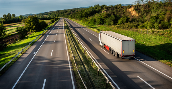 A truck drives on a highway, surrounded by a vibrant green field in the background, showcasing a scenic landscape.