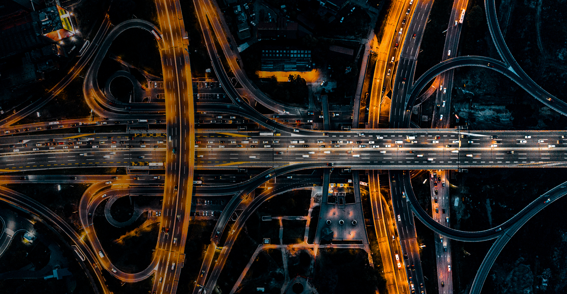 Aerial nighttime view of a large highway interchange with multiple overpasses and looping ramps illuminated by bright orange streetlights, with heavy traffic flowing in several directions.