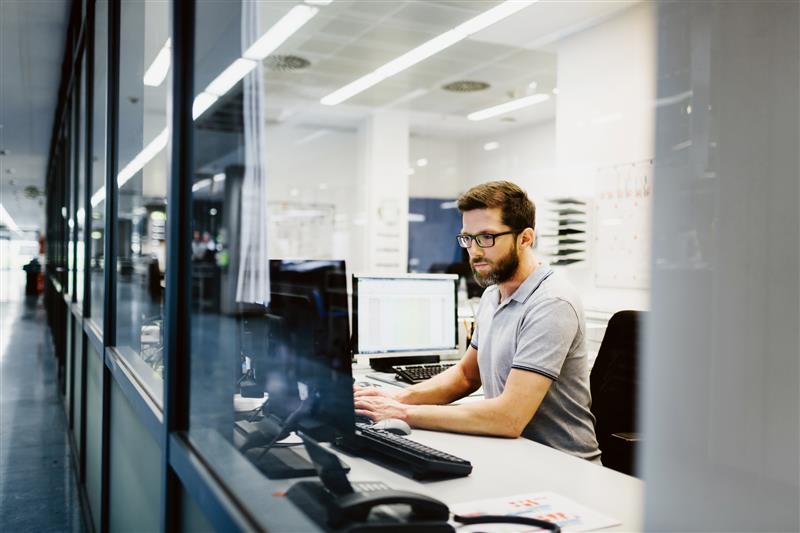 A man wearing glasses is focused on working at his computer, typing and reviewing information on the screen.