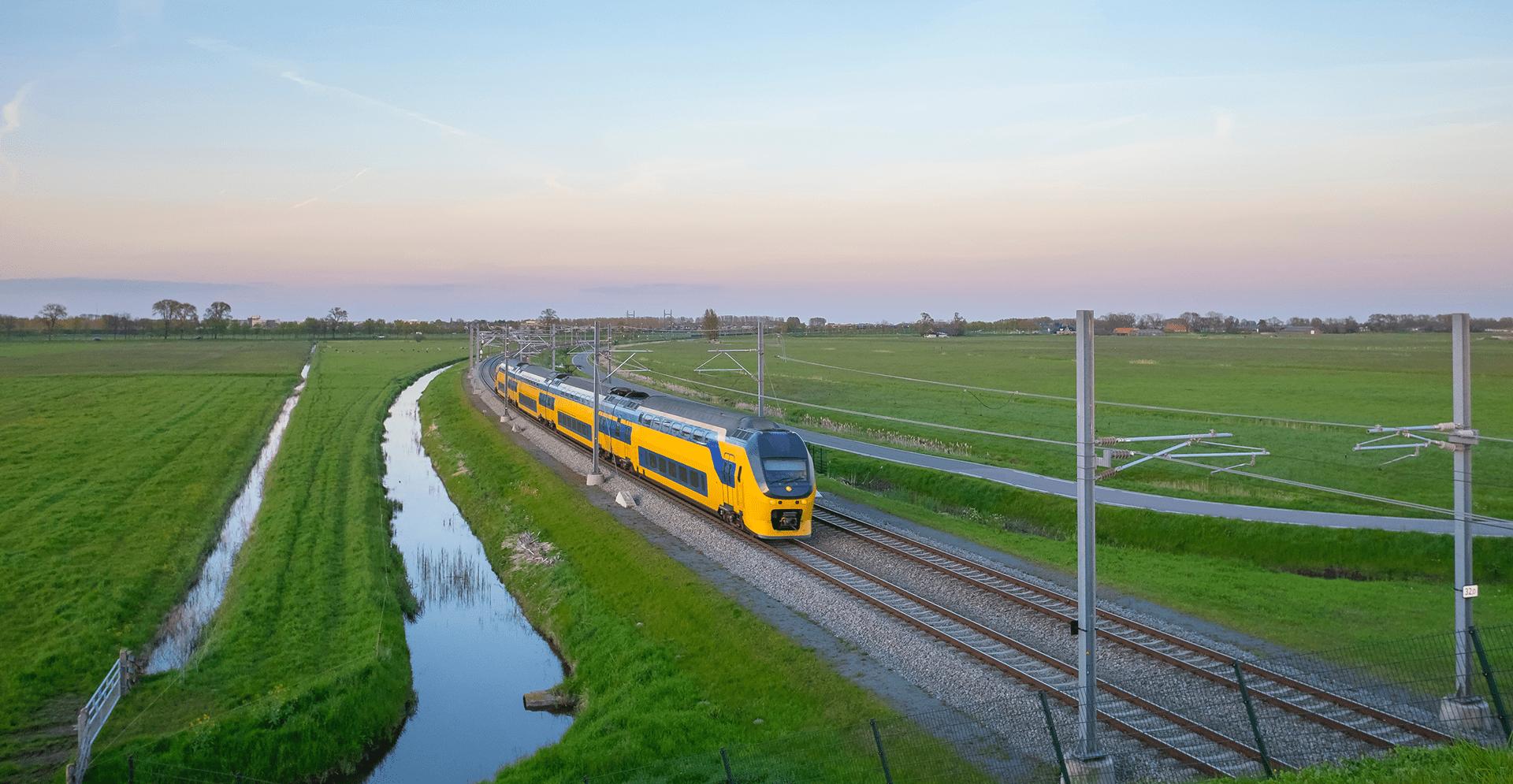 A train moves along the tracks through a scenic rural landscape, surrounded by fields and trees.
