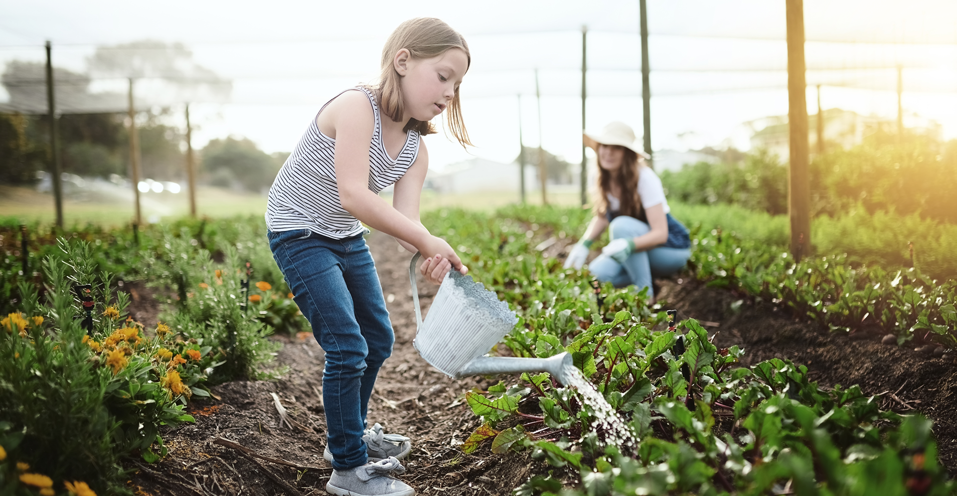 A young girl waters plants in an organic garden, surrounded by vibrant greenery and blooming flowers.