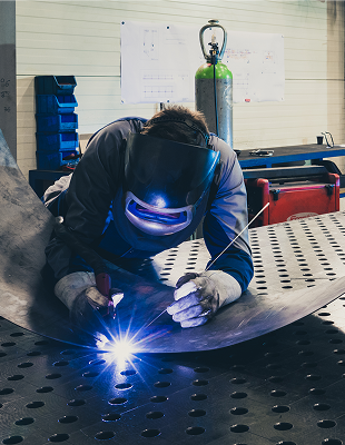 A factory scene showing a welder using a welding torch on steel, surrounded by industrial equipment and safety gear.