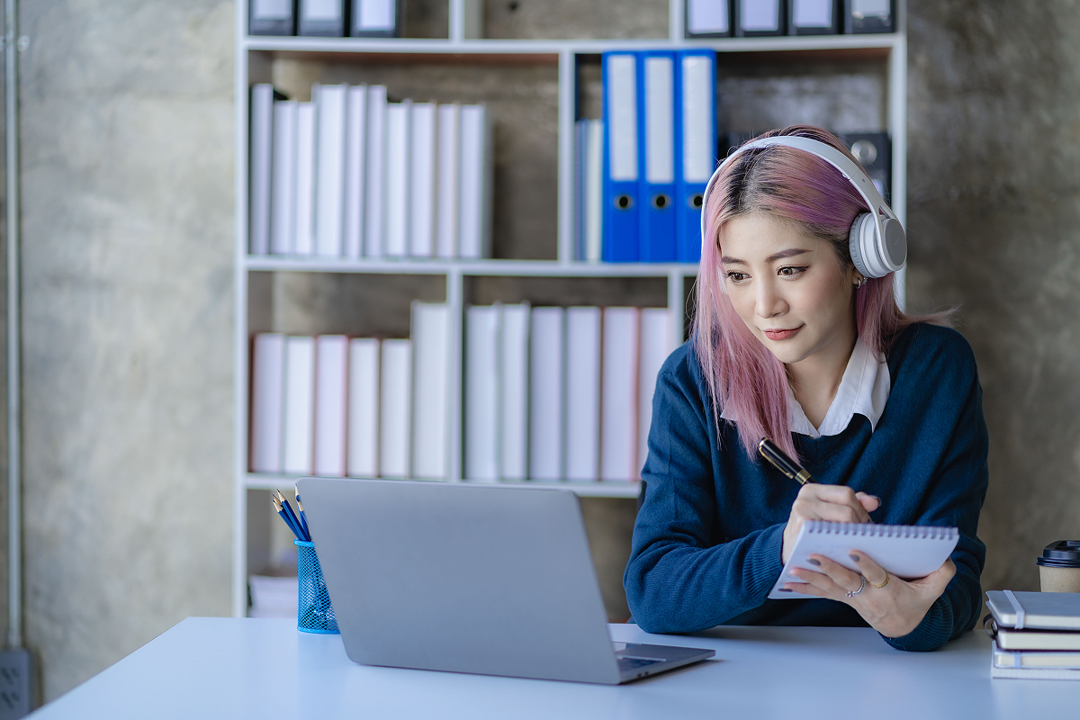 A woman with pink hair wearing headphones, sitting at a desk, focused on her work.
