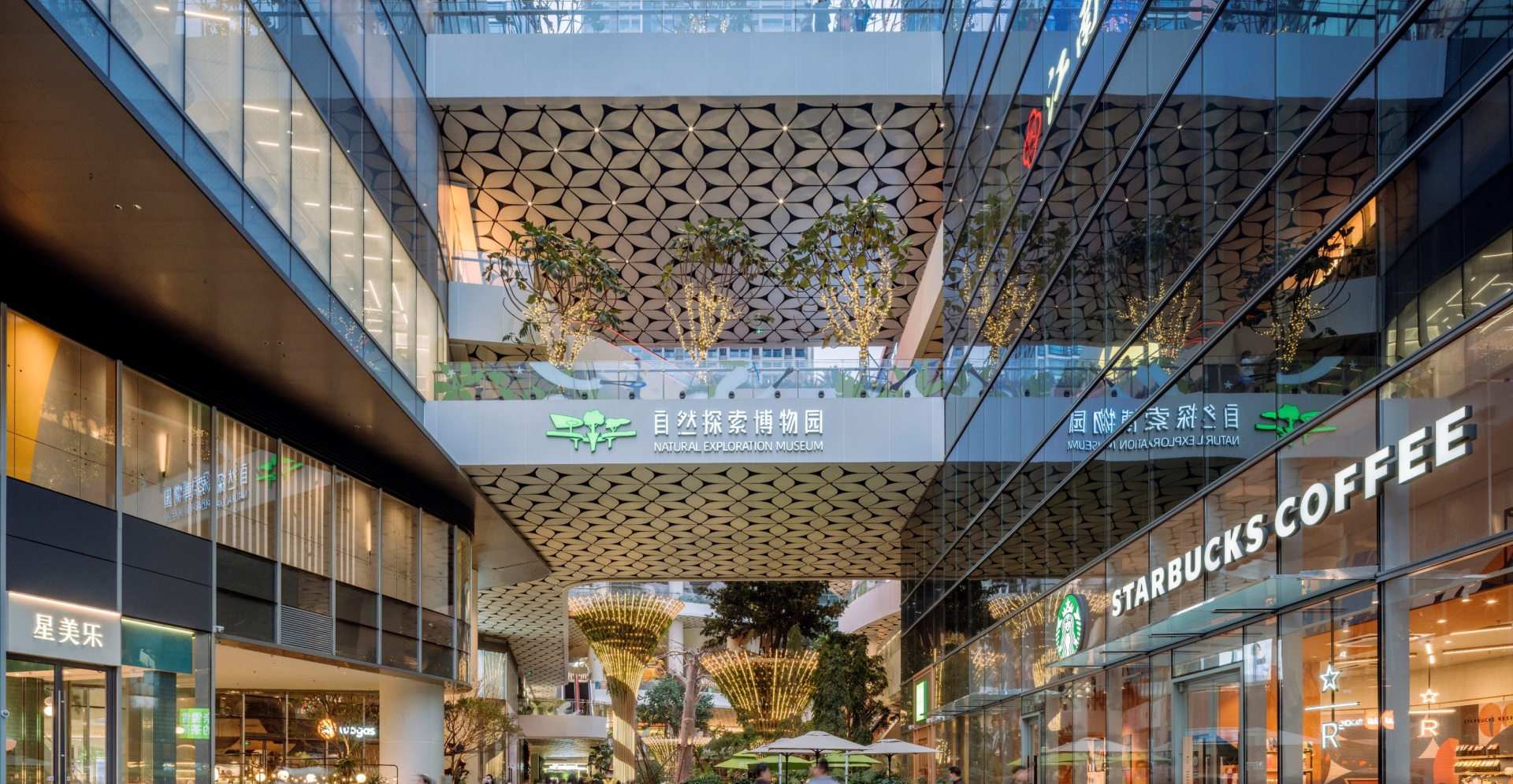 A spacious modern shopping mall interior featuring shops, escalators, and bright lighting with shoppers walking around.