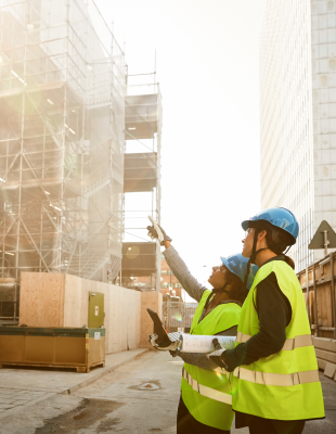 Two construction workers in hard hats stand in front of a partially constructed building, discussing their next steps.
