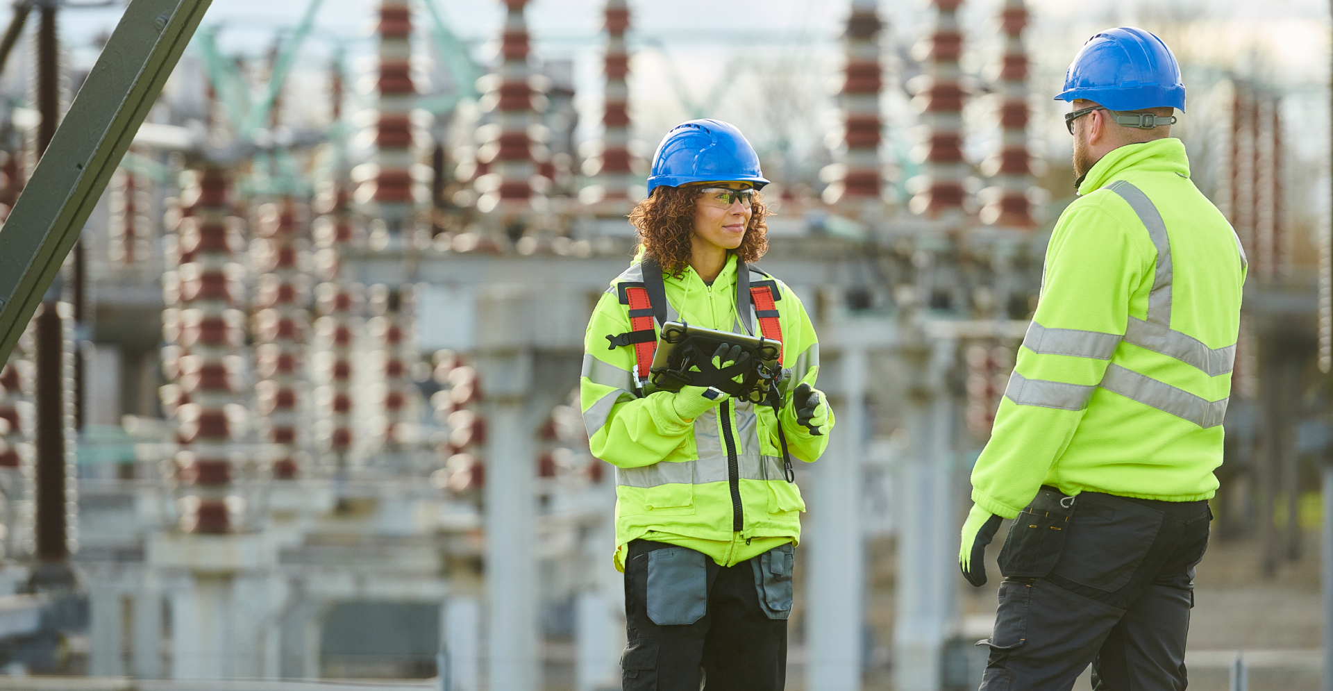 Two individuals in safety vests positioned in front of power lines, emphasizing safety in electrical work.