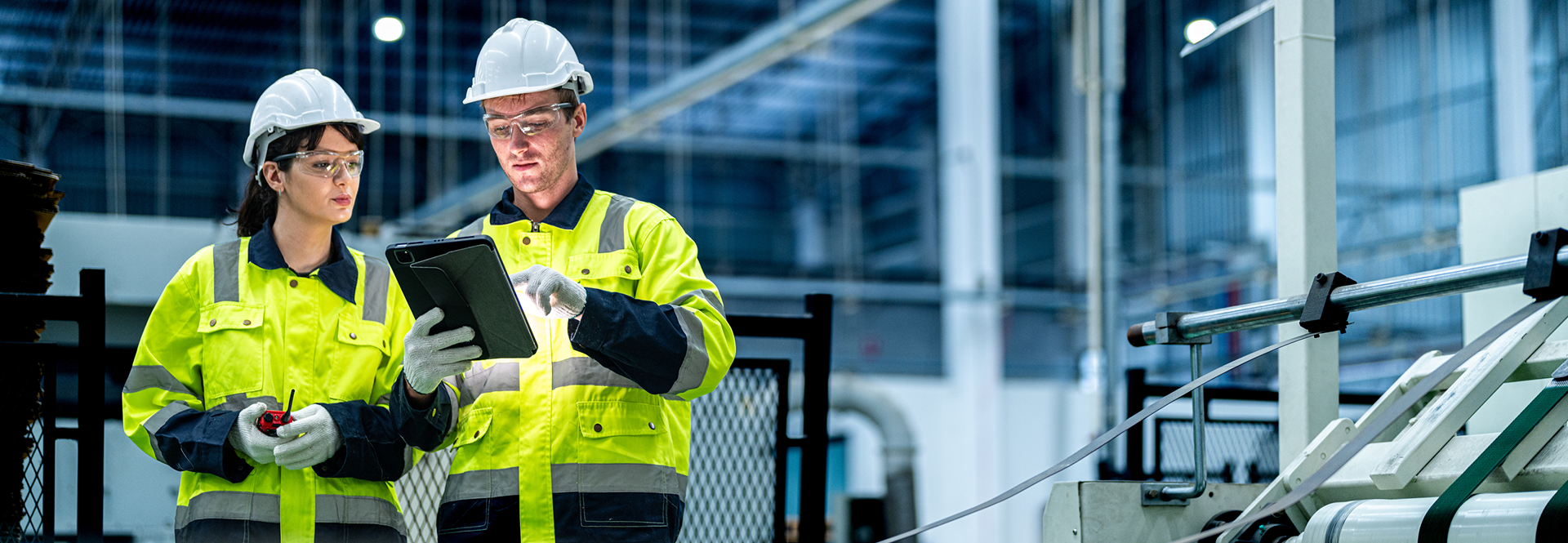 Two safety-equipped workers pose in front of industrial machinery, highlighting safety protocols in a work environment.
